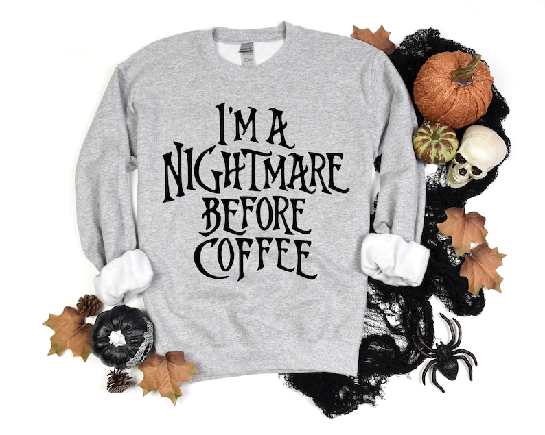 Peut inclure: Un sweat-shirt gris avec le texte "I'm a Nightmare Before Coffee" imprim&eacute; en noir. Le sweat-shirt est photographi&eacute; sur un fond blanc avec des d&eacute;corations d'Halloween, notamment des citrouilles, des toiles d'araign&eacute;es et des feuilles.