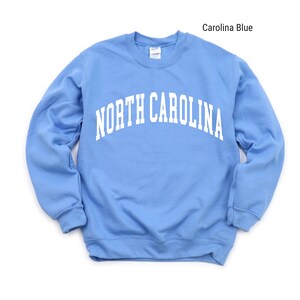 Pode incluir: Uma camisa de moletom azul claro com o texto "NORTH CAROLINA" em branco, em arco na frente.