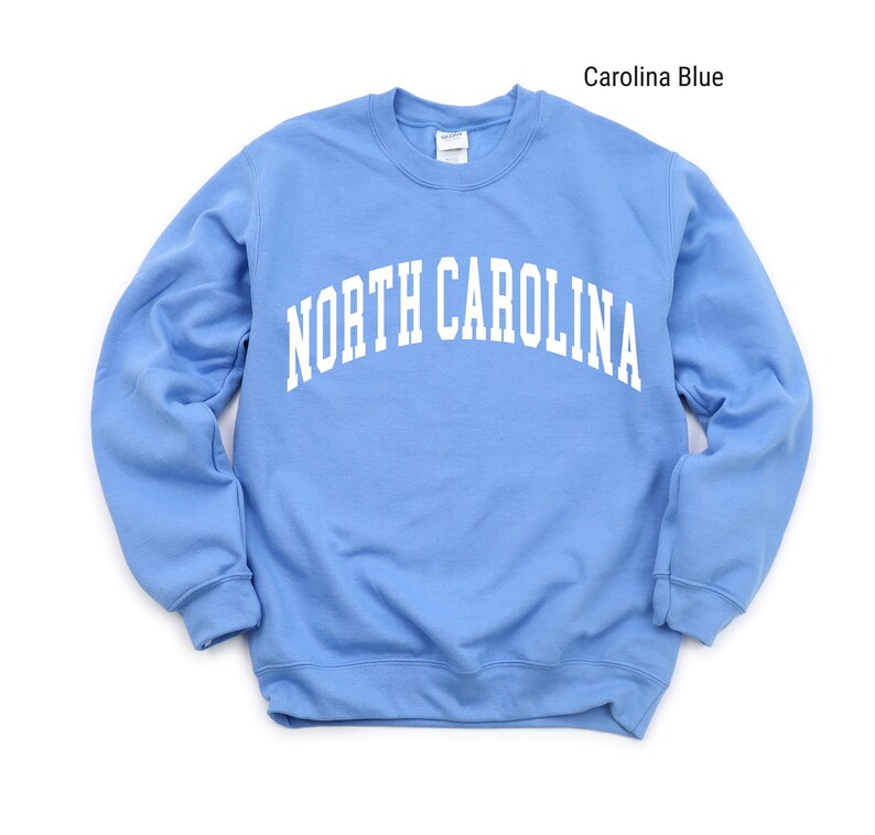 Pode incluir: Uma camisa de moletom azul claro com o texto "NORTH CAROLINA" em branco, em arco na frente.