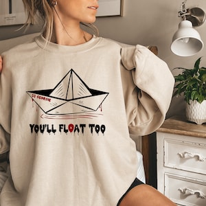 May include: A beige sweatshirt with a black and white illustration of a paper boat with the words "It's Georgie" and "You'll Float Too" printed on it.