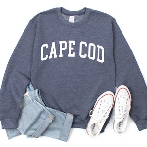 May include: A navy blue sweatshirt with the text "CAPE COD" in white letters. The sweatshirt is folded and laid out on a white background with a pair of white sneakers and blue jeans.