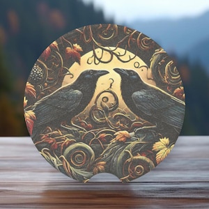 May include: A round car coaster featuring a dark, intricate design of two black ravens perched on branches with a full moon in the background. The design is surrounded by swirling vines and leaves in shades of brown, gold, and green.