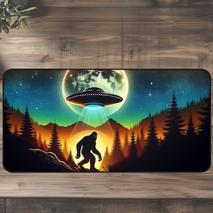 May include: A rectangular mousepad featuring a Bigfoot silhouette in front of a UFO, with a full moon and starry night sky backdrop. The design includes a forest silhouette and a beam of light from the UFO.