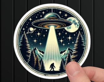 May include: A round sticker featuring a UFO beaming a light down on a Bigfoot silhouette in a forest. The design includes a night sky with stars, a full moon, and a planet. The sticker has a white border.