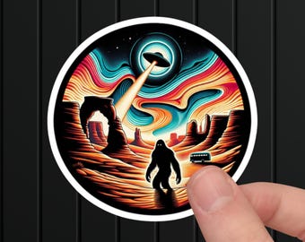 May include: A round sticker featuring a colorful desert landscape with a Bigfoot silhouette, a UFO, and a bus. The sky transitions from dark blue to orange and red. The sticker is held by a finger.