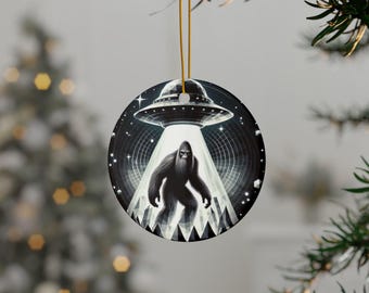May include: A round ceramic Christmas ornament with a black and white illustration of Bigfoot standing in front of a UFO. The UFO is hovering above Bigfoot and shining a beam of light down on him. The background is a dark blue with stars.