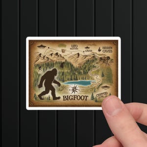 May include: A vintage-style map with a brown background and a silhouette of Bigfoot in the center. The map shows a lake, trails, and the words "UFO Sighting", "Hidden Caves", "Cryptid", and "Bigfoot".