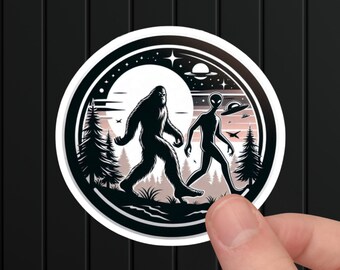 May include: A black and white sticker with a silhouette of Bigfoot and an alien walking side-by-side in a forest. The sticker has a white border and a black background with a moon and stars.