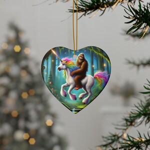May include: Heart-shaped ceramic ornament featuring a whimsical scene of a Bigfoot riding a white unicorn with a rainbow mane and tail. The background depicts a forest with a stream and glowing lights. The ornament hangs from a gold string.