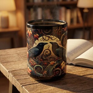 May include: A black ceramic mug featuring a detailed illustration of two dark ravens facing each other amidst swirling autumn leaves, vines, and a pale circular moon. The mug contains a dark liquid with visible steam rising from the top rim.