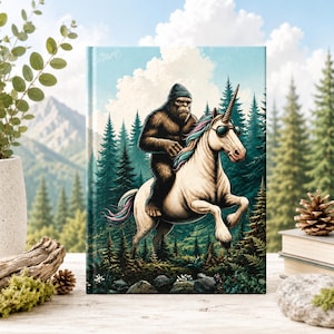 May include: A hardcover journal featuring a graphic illustration of a brown Bigfoot wearing a dark beanie, riding a white unicorn with a multicolored mane and tail. The unicorn wears black sunglasses. The background depicts a forest and mountains.