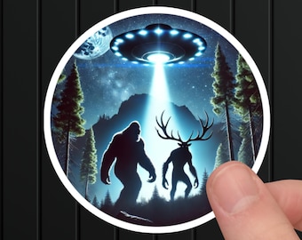 May include: A round sticker with a blue and white UFO hovering over a dark forest scene.  Two shadowy figures, one resembling Bigfoot and the other a deer-like creature, stand in the forest.  The UFO is emitting a bright beam of light.