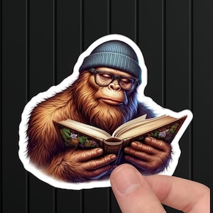 May include: A brown and orange Bigfoot wearing a blue beanie and glasses is reading a book with colorful illustrations.