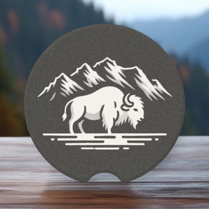 May include: A gray round coaster with a white silhouette of a bison standing in front of a mountain range.