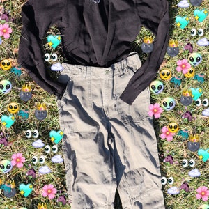 Pode incluir: Um su&eacute;ter preto de gola alta com um colar de corrente prateada, combinado com cal&ccedil;as cargo cinza claro. A roupa est&aacute; disposta em um leito de grama verde com gr&aacute;ficos coloridos que se assemelham a emojis.