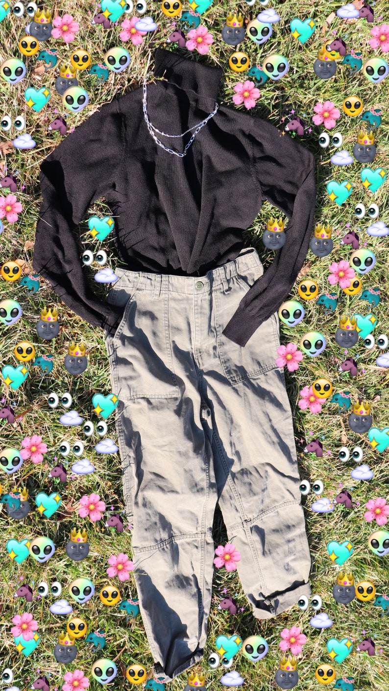 Pode incluir: Um su&eacute;ter preto de gola alta com um colar de corrente prateada, combinado com cal&ccedil;as cargo cinza claro. A roupa est&aacute; disposta em um leito de grama verde com gr&aacute;ficos coloridos que se assemelham a emojis.