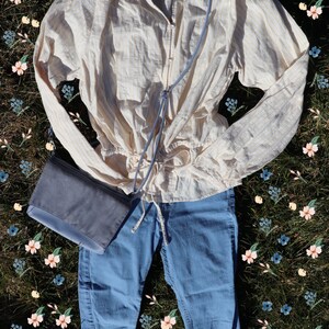May include: A light yellow and white striped blouse with a drawstring waist, blue jeans, and a gray crossbody bag. The outfit is laid out on a bed of green grass with a scattering of pink and blue flowers.