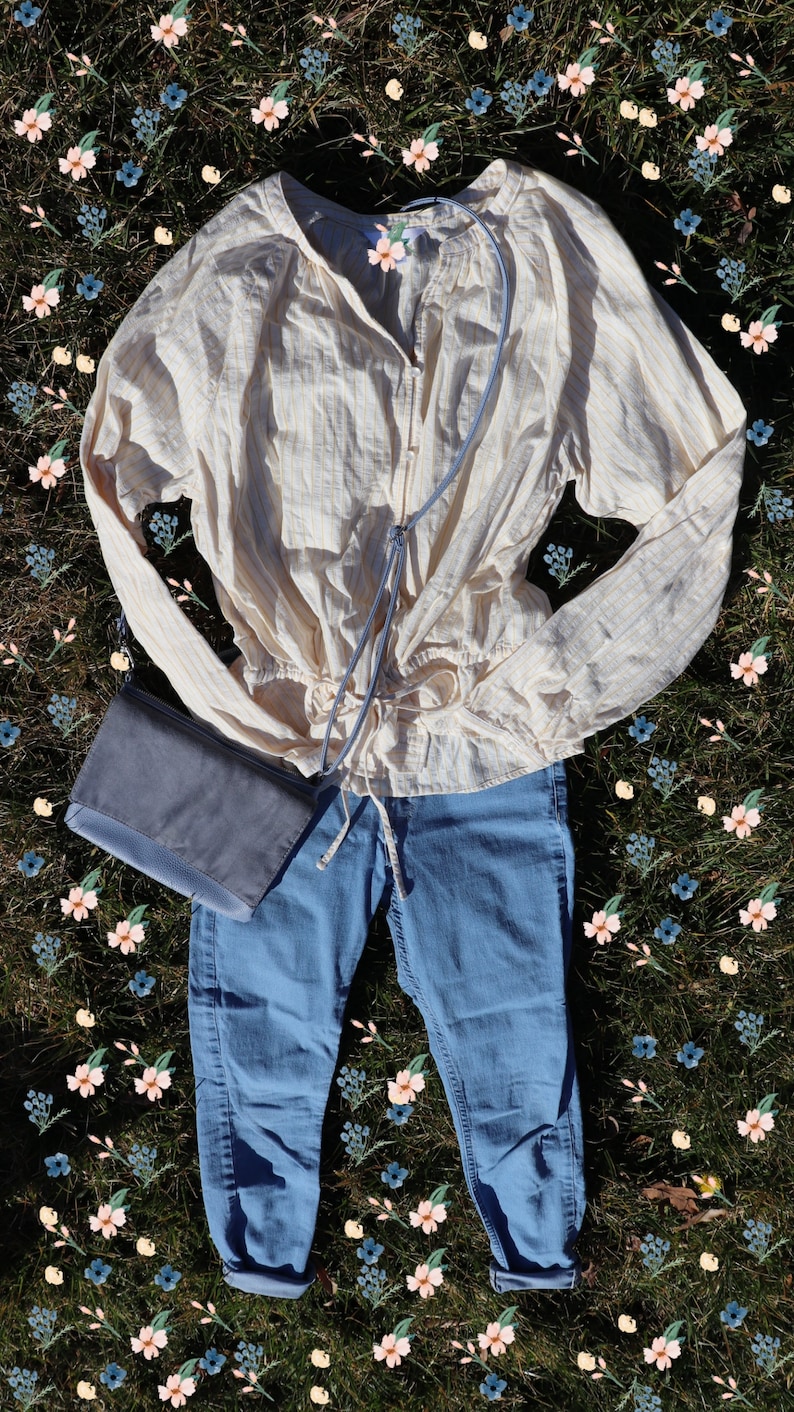 May include: A light yellow and white striped blouse with a drawstring waist, blue jeans, and a gray crossbody bag. The outfit is laid out on a bed of green grass with a scattering of pink and blue flowers.