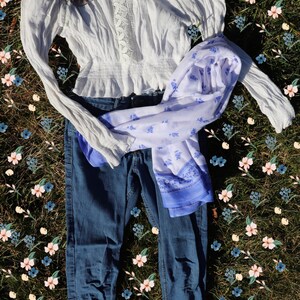 May include: A white off-the-shoulder blouse with lace detailing, blue jeans, and a white and blue patterned scarf laid out on a bed of green grass with pink and blue flowers.