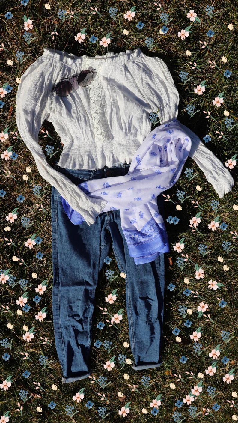 May include: A white off-the-shoulder blouse with lace detailing, blue jeans, and a white and blue patterned scarf laid out on a bed of green grass with pink and blue flowers.