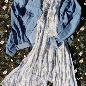 May include: A white floral dress with a light blue denim jacket on top. The dress and jacket are laid out on a bed of green grass with pink and blue flowers.