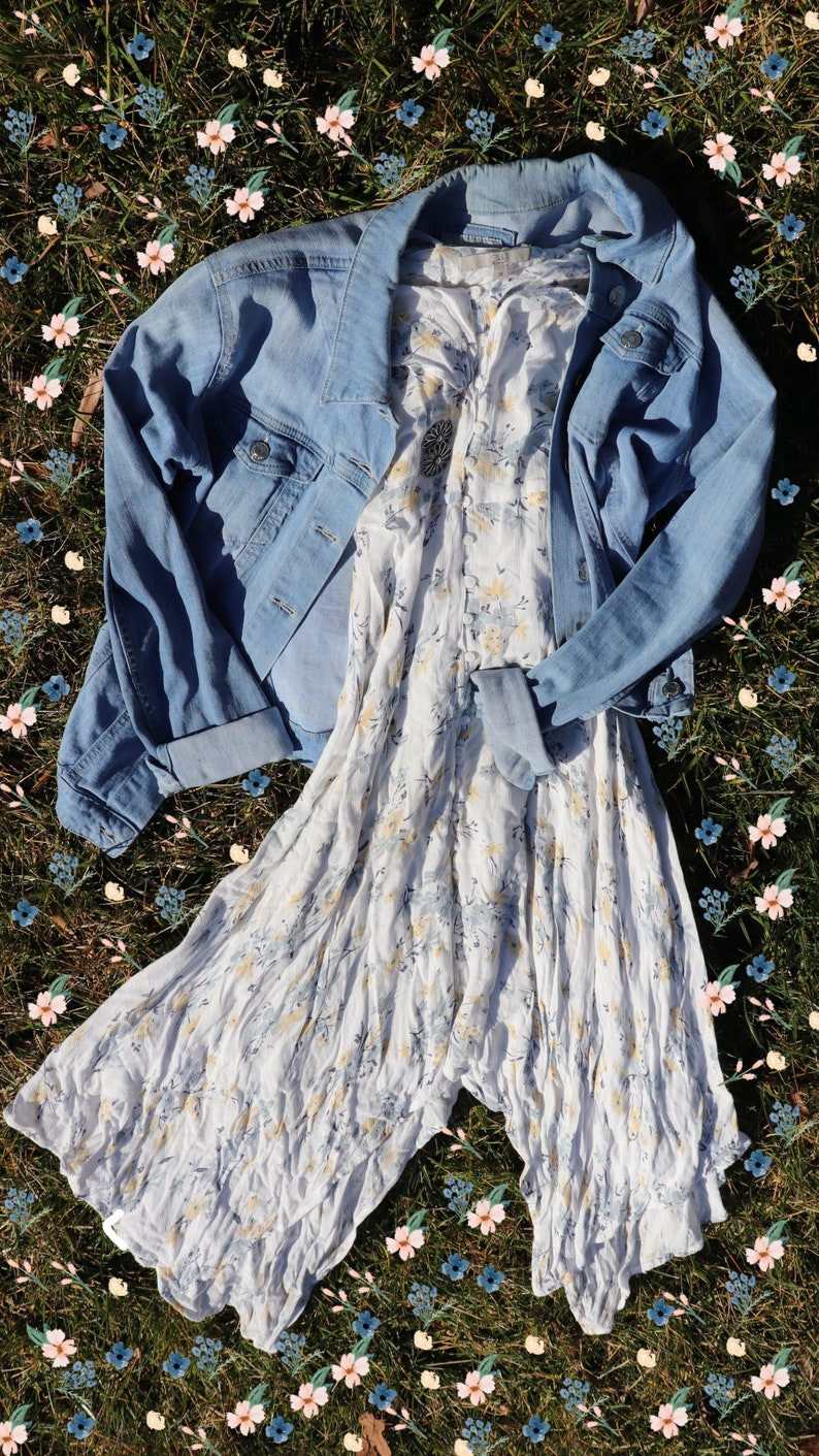 May include: A white floral dress with a light blue denim jacket on top. The dress and jacket are laid out on a bed of green grass with pink and blue flowers.