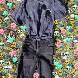 Pode incluir: Um gorro preto, uma camiseta cinza escura com o texto "The Bahamas" e um gr&aacute;fico, e cal&ccedil;as jeans pretas com joelhos desgastados s&atilde;o colocados em uma cama de grama verde com adesivos de emoji coloridos.