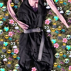 Pode incluir: Um macac&atilde;o preto com uma camisa rosa de manga comprida sobreposta. O macac&atilde;o tem um cinto largo cinza e a camisa tem uma textura canelada. O conjunto est&aacute; colocado sobre um leito de grama verde com gr&aacute;ficos de desenhos animados coloridos.