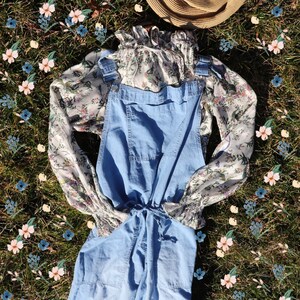 May include: A light blue denim overall with a floral print blouse and a straw hat on a bed of green grass with pink and blue flowers.