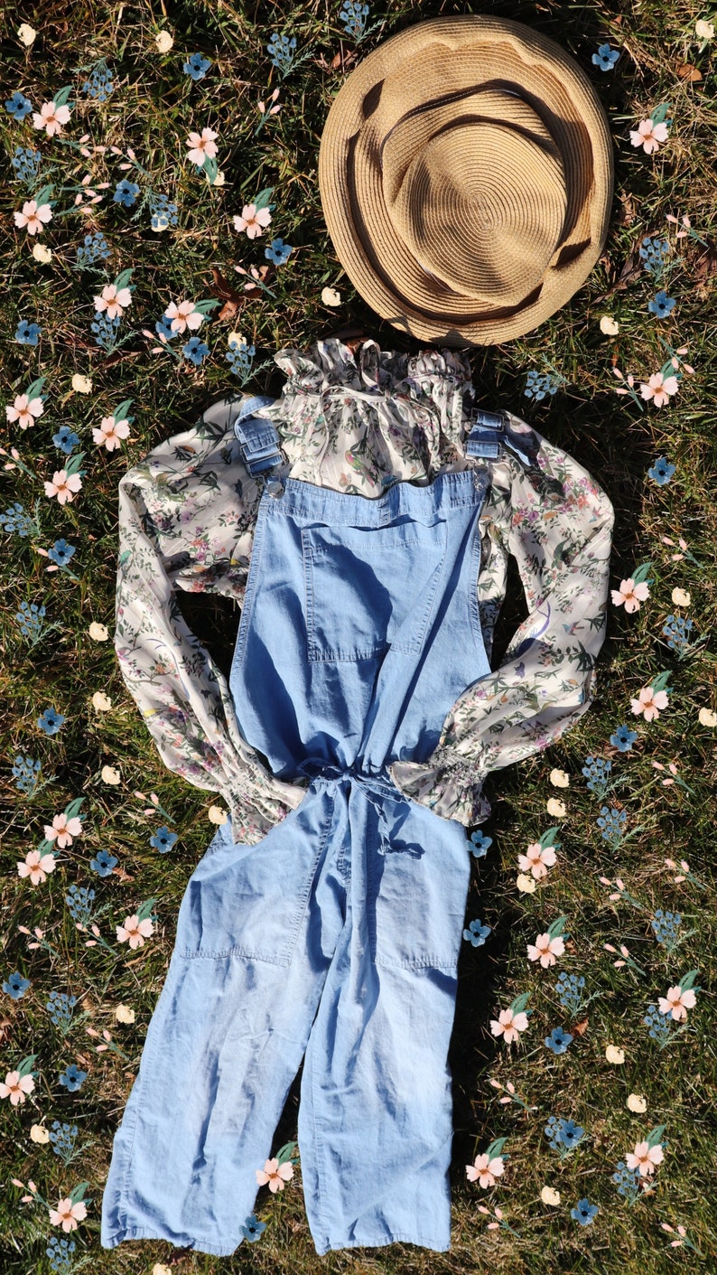 May include: A light blue denim overall with a floral print blouse and a straw hat on a bed of green grass with pink and blue flowers.
