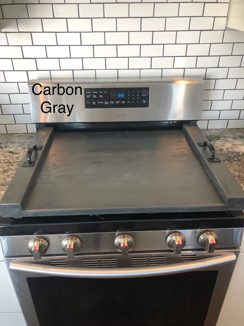 May include: A stainless steel stovetop with a black, wooden, rectangular stovetop cover. The stovetop has four red knobs and a Samsung logo. The stovetop cover has two black handles. The stovetop is against a white tiled wall.