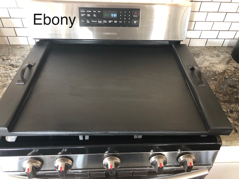 May include: Black stovetop griddle with two handles. The griddle is made of cast iron and has a black finish. The griddle is sitting on top of a stainless steel stove.