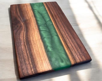 May include: A rectangular wooden cutting board with a green epoxy resin inlay. The board has a natural wood grain pattern and is perfect for serving cheese, charcuterie, or other appetizers.