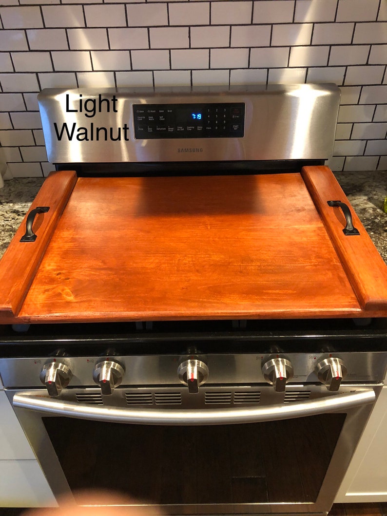 May include: A light walnut wood stovetop cover with handles. The cover is placed on top of a stainless steel stove with a Samsung logo. The stove has four burners and an oven.