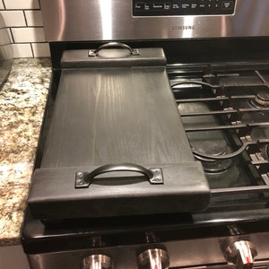 May include: Black wooden stovetop cover with two metal handles. The cover is sitting on a stainless steel stovetop with a digital clock display that reads 9:03.
