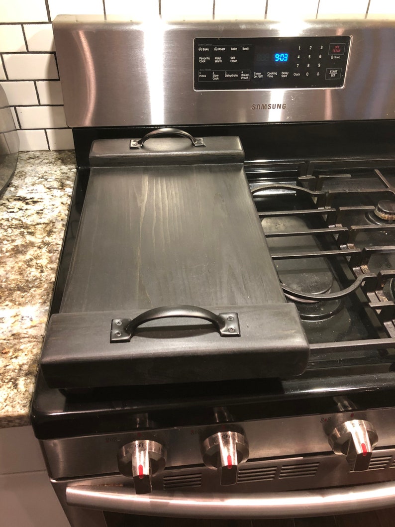 May include: Black wooden stovetop cover with two metal handles. The cover is sitting on a stainless steel stovetop with a digital clock display that reads 9:03.