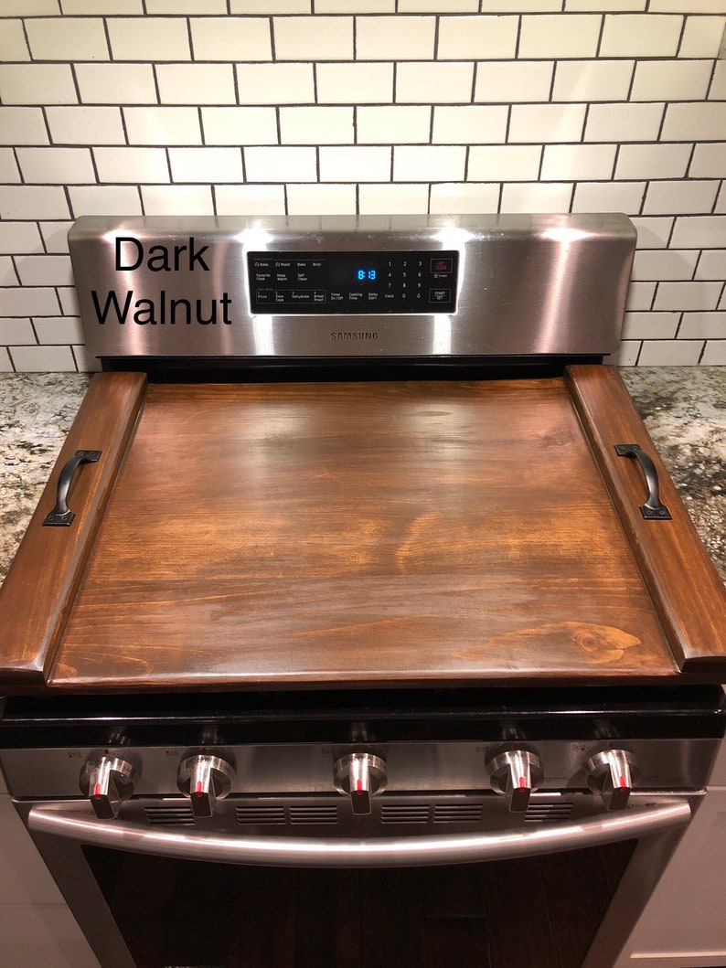 May include: Dark walnut wood stovetop cover with handles. The cover is placed on top of a stainless steel stove with four burner knobs and an oven door.
