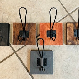 May include: Five wooden wall hooks with black metal hardware. The square wooden bases are stained in various colors: black, brown, orange, and gray. Each hook has a looped top and a lower hook.