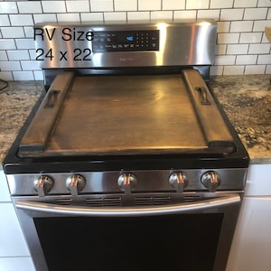 May include: A stainless steel stovetop with a wooden cutting board on top. The stovetop has four burners and an oven. The cutting board is 24 inches by 22 inches. The stovetop is labeled "Samsung".