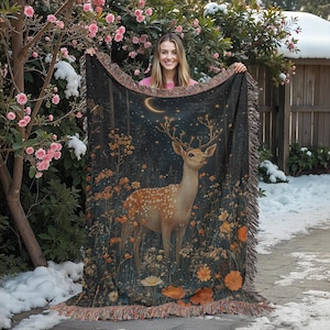 May include: A woven throw blanket with a dark background featuring a deer standing in a field of flowers under a crescent moon. The deer is brown and white with a spotted pattern. The flowers are in shades of yellow, orange, and red.