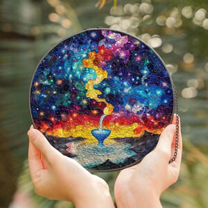 May include: A round stained glass window with a colorful mosaic design depicting a starry night sky with a waterfall pouring into a pool of water. The window is framed in a black metal hoop and has a chain attached.