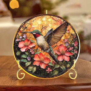 Op de afbeelding: Een ronde decoratieve glas-in-lood paneel met een kolibrie in vlucht, omringd door rode en roze bloemen en groen blad. Het paneel is gevat in een goudkleurig metalen frame en wordt ondersteund door een gouden standaard. De achtergrond heeft een mozaïekachtig uiterlijk.