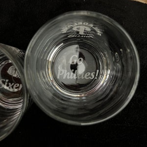 May include: Two clear glass tumblers with etched designs. One tumbler has the text "Go Phillies!" etched on the bottom. The other tumbler has the text "Go Sixers!" etched on the bottom.