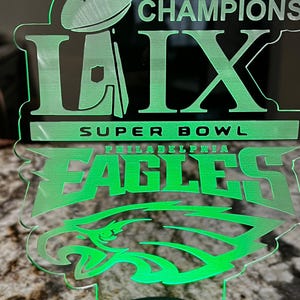 May include: Green illuminated acrylic sign with the words "Champions LIX Super Bowl Philadelphia Eagles" and a green silhouette of an eagle's head.
