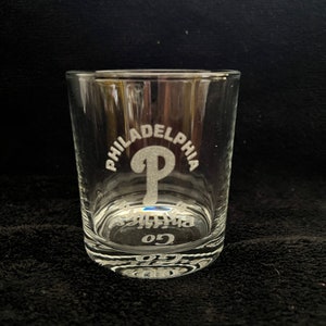 May include: A clear glass tumbler with an etched design of the Philadelphia Phillies logo and the word "Philadelphia". The bottom of the glass has the number "05" etched into it.