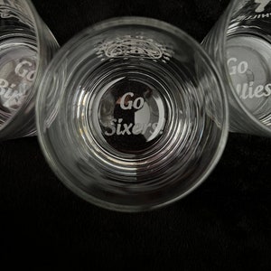May include: Three clear glass tumblers with the words "Go Sixers!" etched on the bottom. The glasses are stacked on top of each other.
