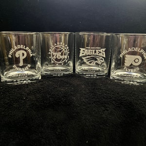 May include: Four clear glass tumblers with etched designs of Philadelphia sports teams: Phillies, 76ers, Eagles, and Flyers.
