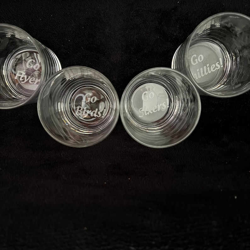 May include: Four clear glass tumblers with etched designs. Each tumbler features a different sports team logo with the text "Go Flyers", "Go Birds", "Go Sixers", and "Go Phillies!"