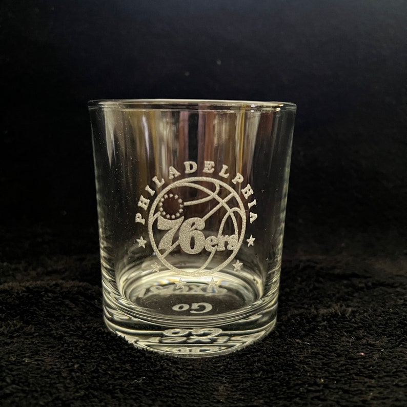 May include: A clear glass tumbler with an etched design of the Philadelphia 76ers logo. The logo features a basketball with the team's name and number "76ers" inside a circle.