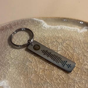 Könnte beinhalten: Ein rechteckiger, silberfarbener Schlüsselanhänger mit Spotify-Code und -Logo. Er ist an einem silbernen Schlüsselring befestigt. Der Hintergrund ist eine strukturierte, cremefarbene Oberfläche.